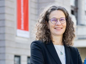 Es ist eine Frau mit einem schwarzen Blazer und einem weißen T-Shirt vor einem grauen Haus zu sehen.