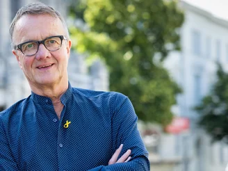 Auf dem Bild ist ein Mann in blauem Blazer und mit blauem Hemd zu sehen, der vor Bäumen steht.
