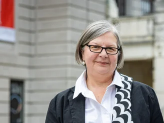 Es ist eine Frau mit einer schwarzen Jacke und einer weißen Bluse vor einem grauen Haus zu sehen.