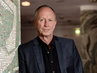 Auf dem Bild ist ein Mann in einem schwarzen Anzug zu sehen, der gegen eine Wand aus Mosaik-Steinen gelehnt ist.