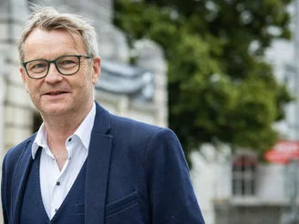 Es ist ein Mann in blauem Anzug zu sehen, der vor einem grauen Haus und Bäumen steht.