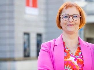 Auf dem Bild ist eine Frau in einem pinken Blazer zu sehen, die vor einem grauen Haus steht.