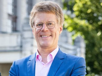 Es ist ein Mann in einem hellblauen Blazer und einem rosa Hemd vor einem grauen Haus und Bäumen zu sehen.